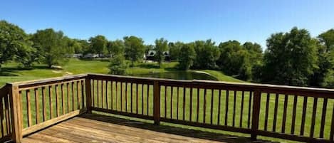 A wooden deck with a railing, overlooking a green landscape with trees and a clear blue sky.