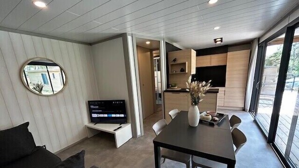 Una sala de estar moderna con televisor de pantalla plana, una mesa de comedor con sillas, un sofá y un ventanal con vistas al exterior.