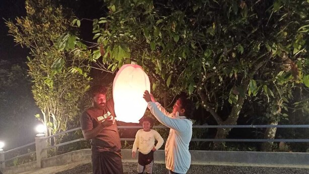 A group of people outdoors at night, with a large illuminated object in the center. - 	“Your Family’s Perfect Getaway by the Riverside!” (Mankulam)