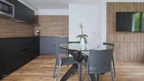 Uma cozinha moderna com armários escuros, piso de madeira, mesa de jantar de vidro e duas cadeiras, além de um vaso com planta.