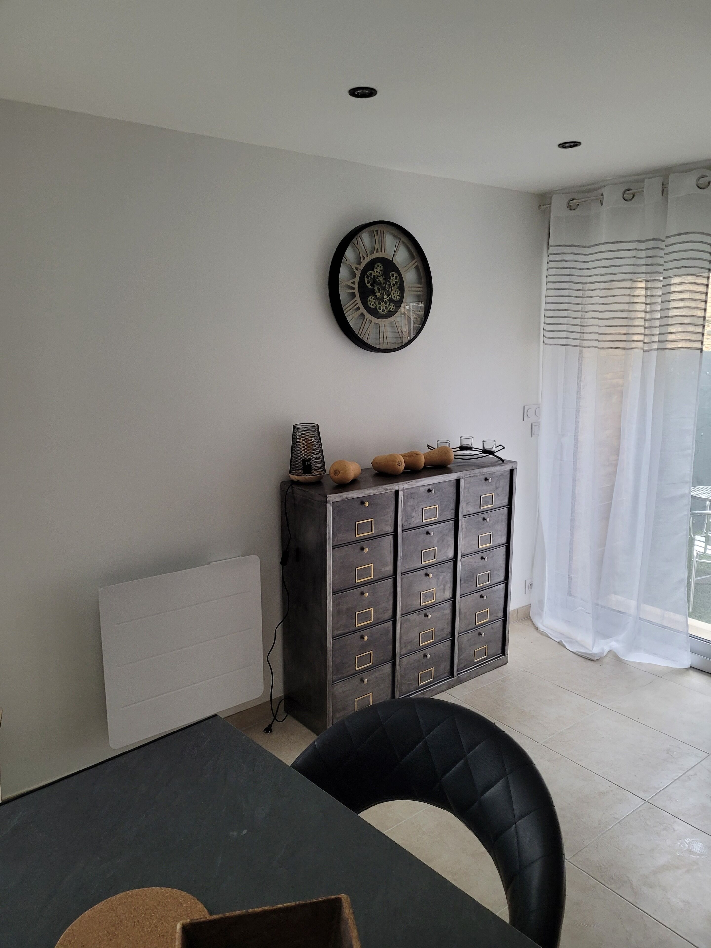 A dining area with a table, chairs, a cabinet with decorative items, a wall clock, and a window with curtains.