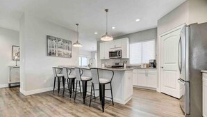 A modern kitchen with a central island, bar stools, and a refrigerator. - Rooftop Patio, Hot Tub, EV charging, Mtn views, close to AFA! (Palmer Lake)