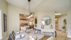 A dining area with a wooden table set for six, surrounded by chairs. A chandelier hangs above, and a large mirror is on the wall.
