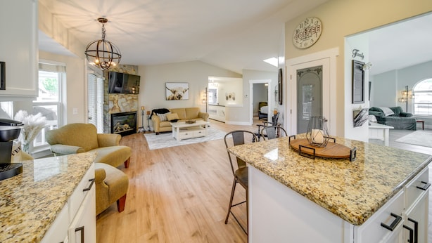 A spacious kitchen with a large island, wooden flooring, and a chandelier. There is a dining area with a table and chairs, and a living room with a sofa and a fireplace.