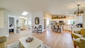 A spacious living room with a wooden floor, a dining area with a high table, and a kitchen with white cabinets and a farmhouse sign.