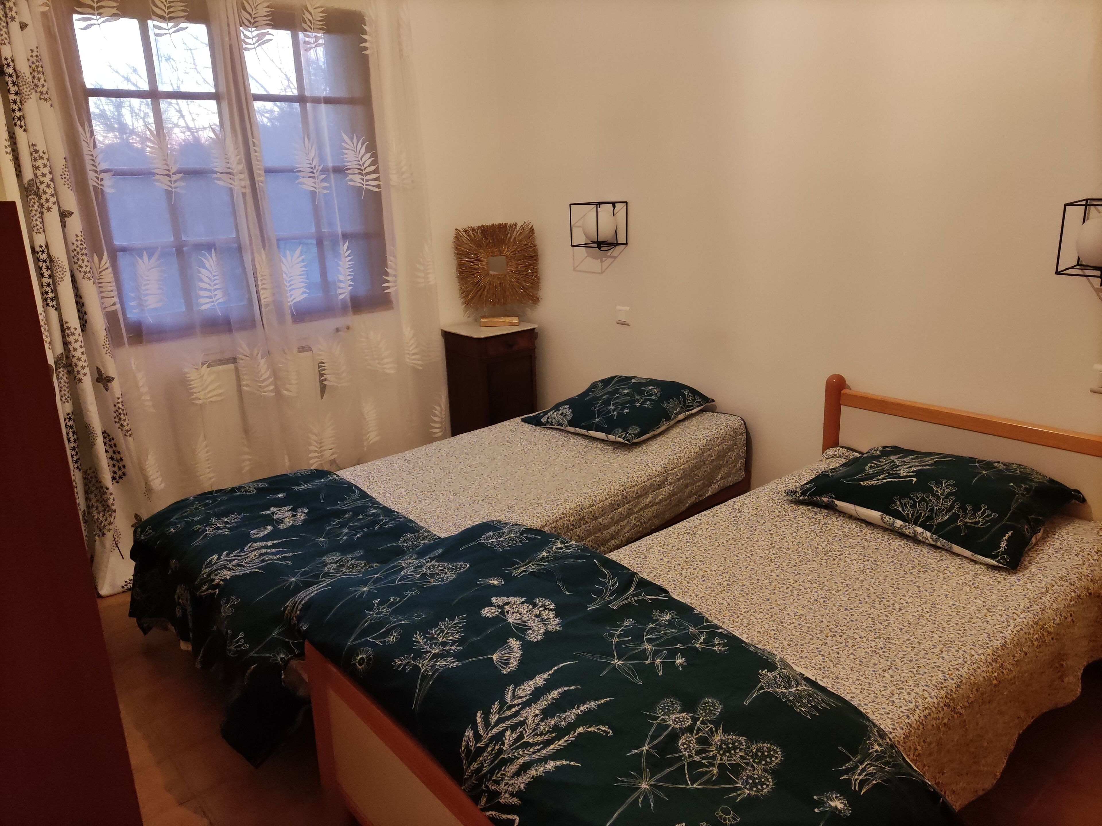 Een slaapkamer met twee eenpersoonsbedden, een raam met dunne gordijnen, een klein nachtkastje en wandlampen.