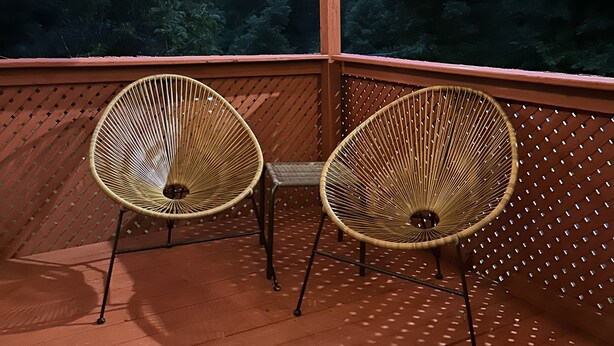 Una terrazza in legno con due sedie in vimini e una panca, con vista su una zona boschiva.