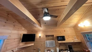 A wooden cabin interior with a ceiling fan, mounted TV, and a kitchenette area.