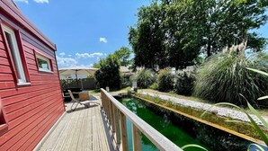 A wooden deck with a pool, surrounded by greenery and a red building. - Charming family home- natural pool (Gujan-Mestras)