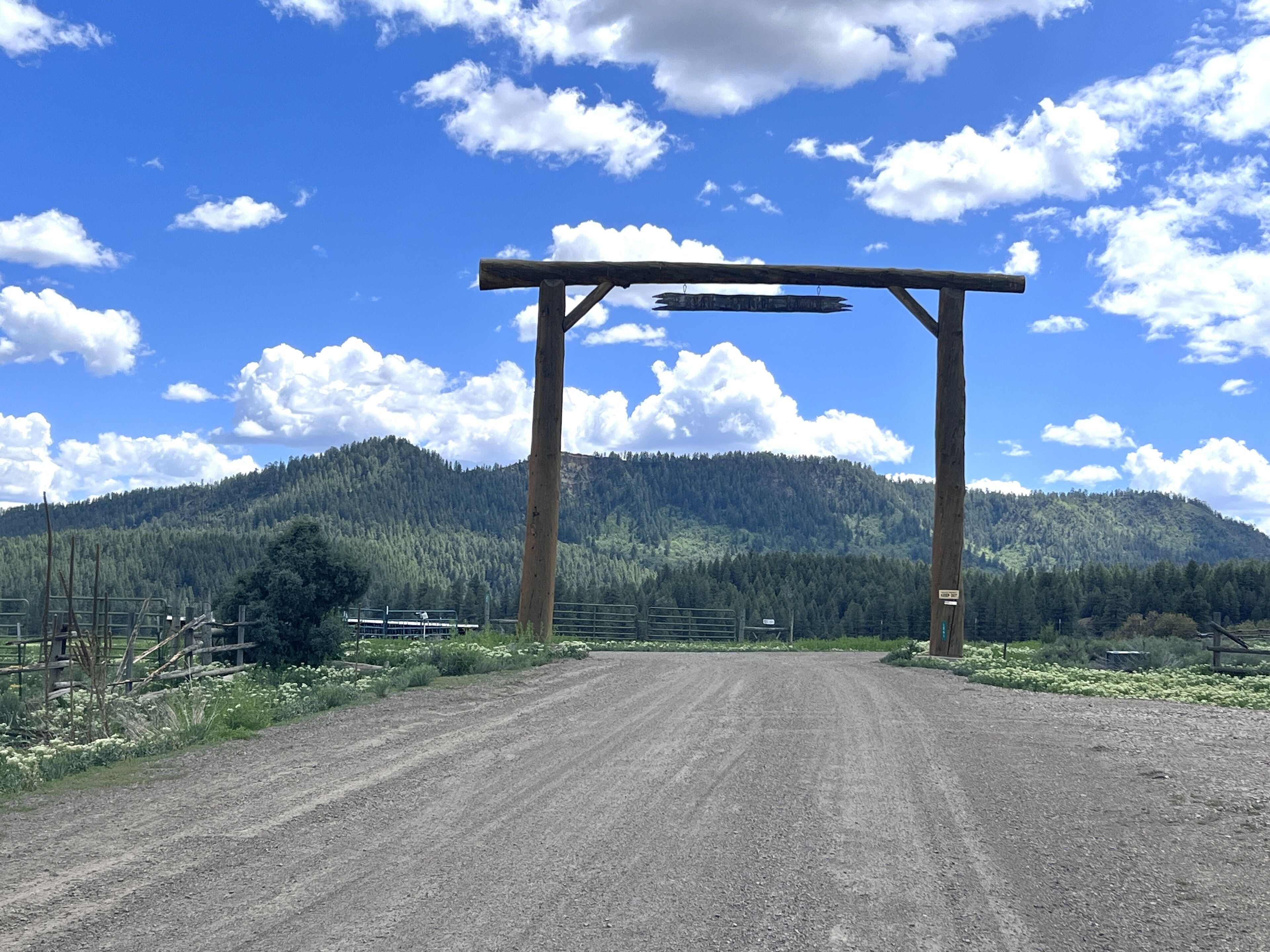 Ein Feldweg führt zu einem hölzernen Torbogen, dessen Hintergrund von einem bewaldeten Berg gebildet wird.