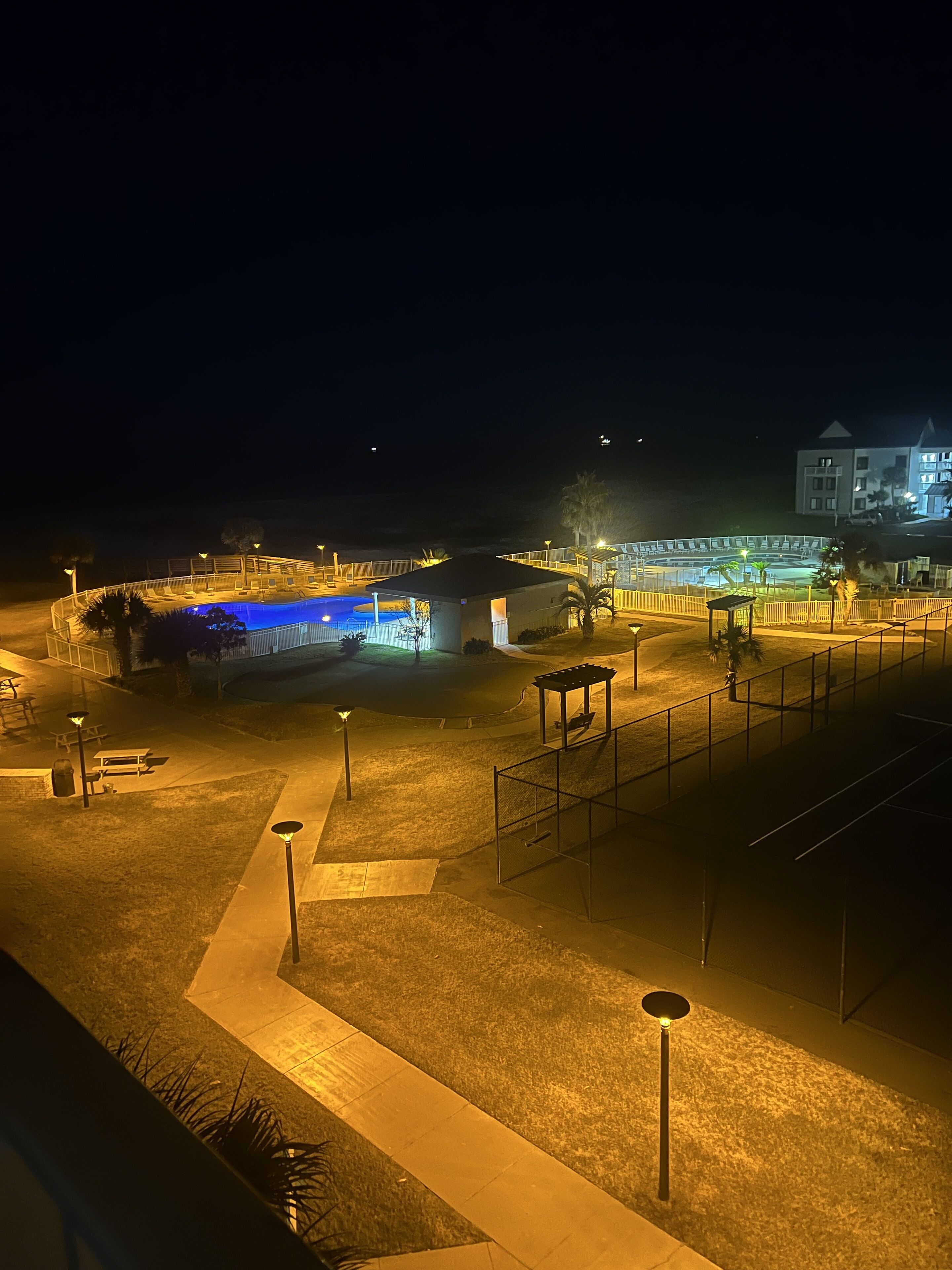 A nighttime view of a pool area with a covered seating area, a building with a lighted facade, and a fenced pool.