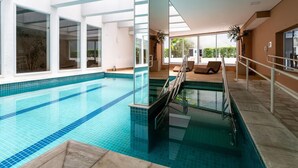 Ein moderner, überdachter Schwimmbereich mit klarem, blauem Wasser, Glaswänden und einem sonnendurchfluteten Liegebereich.