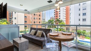 Ein modernes Wohnzimmer mit Sofa, Couchtisch und klaren Stühlen. Ein Holzesstisch mit Glasplatte ist zu sehen. Große Fenster bieten Blick auf Gebäude und einen Swimmingpool.