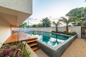 Een modern buitenzwembad met een houten terras, omgeven door planten en een betonnen rand.