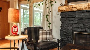 A cozy living room with a stone fireplace, a leather chair with a plaid pillow, a small table with a red lantern, and a view of the outdoors through a window. - Scenic Kettle River Cabin - Hike, Sauna, Kayaks & More-Your Next Retreat Awaits! (Willow River)