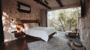 Une chambre avec un mur en pierre, un lit en bois, une table de chevet, une chaise et une grande fenêtre donnant sur des arbres.