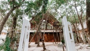 Une construction rustique en bois, entourée d’arbres, avec un portail blanc en premier plan.