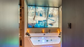 Une salle de bain moderne avec une grande baignoire, un téléviseur fixé au mur qui diffuse un paysage enneigé, et deux bougies allumées sur le meuble-lavabo.