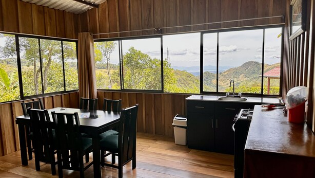 A room with a wooden interior, a dining table with chairs, a kitchen area with a sink, and large windows offering a view of greenery and hills. - Campo Azul #3 - Monteverde (Provincia de Puntarenas)