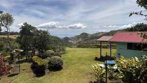A green lawn with a small building with a red roof, surrounded by trees and hills in the background. - Campo Azul #3 - Monteverde (Provincia de Puntarenas)
