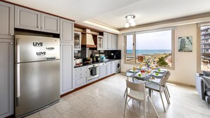 A modern kitchen with a view of the sea, a dining area with a table set for two, and a large window overlooking the beach. - Mackenzie Eftyhia Beachfront Suite (Larnaca)