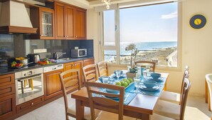 A kitchen with wooden cabinets, a microwave, and a stove. There is a dining table with chairs and a view of the beach through large windows. - Mackenzie Eftyhia Beachfront Suite (Larnaca)