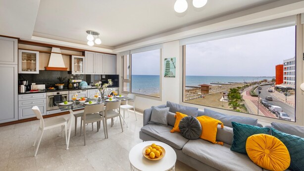 A modern kitchen with a dining area, large windows with a sea view, and a living room with a sofa and cushions. - Mackenzie Eftyhia Beachfront Suite (Larnaca)