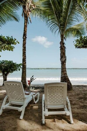 Two white lounge chairs under palm trees by the beach with a view of the ocean.