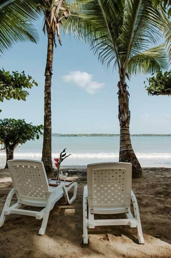 Two white lounge chairs under palm trees by the beach with a view of the ocean.