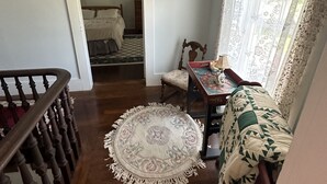 Una habitación con una escalera de madera, un tapete redondo, un sofá con funda estampada, una mesita con una lámpara y una ventana con cortinas de encaje.