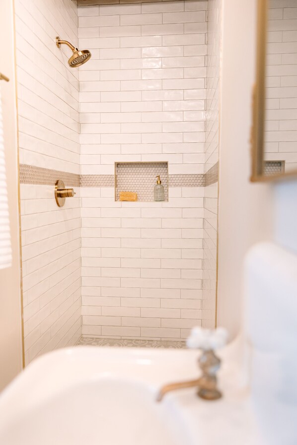Baño con azulejos blancos tipo metro, cabezal de ducha dorado, dispensador de jabón de vidrio y bañera blanca.