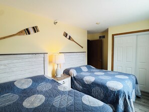 A hotel room with two beds, a nightstand, a lamp, and a wooden ceiling decoration. - Wake, Swim, Repeat—Poolview Fun | 8 ppl | C4 (Put-in-Bay)
