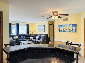 A living room with a black leather sofa, a dining table with chairs, a ceiling fan, and a painting of sailboats on the wall.