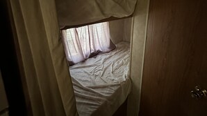 Een kleine, zwak verlichte kamer met een enkel bed, een houten deur en een gedeeltelijk dichtgetrokken gordijn.