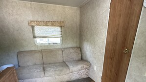 Een compact interieur van een camper met een bank, een raam met zonwering, een houten deur en een betegelde vloer.