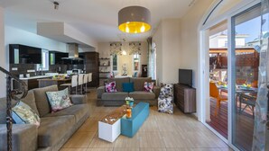 Un salon moderne avec un canapé, une table basse et un téléviseur. On y trouve également un coin cuisine avec des meubles sombres et un espace repas visible à travers une porte vitrée.