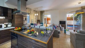 Une cuisine moderne avec un îlot central, un coin repas avec une table et des chaises, et un salon avec un canapé et un téléviseur.