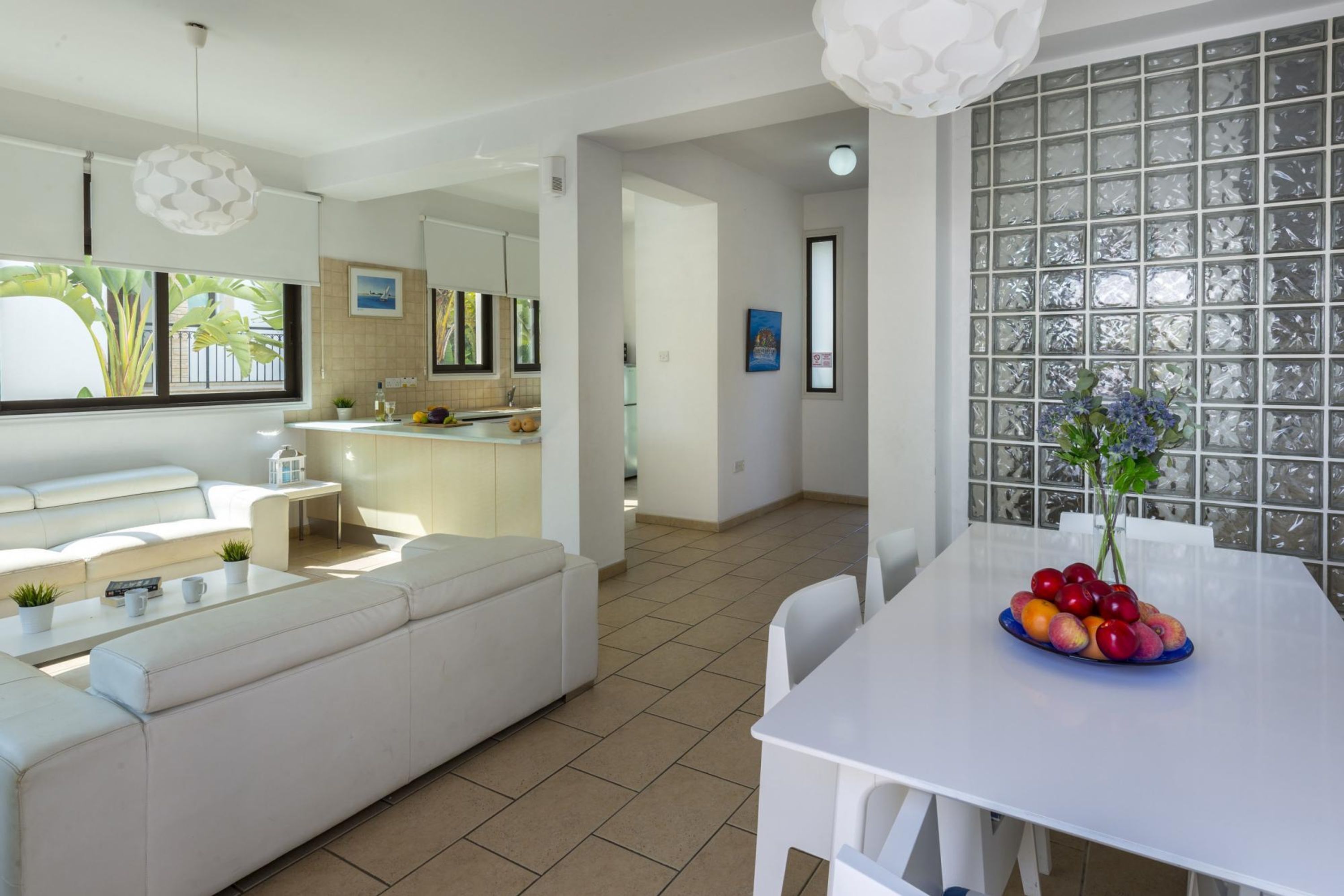 A modern living room with a white sofa, a dining table with fruit, and a kitchen in the background.