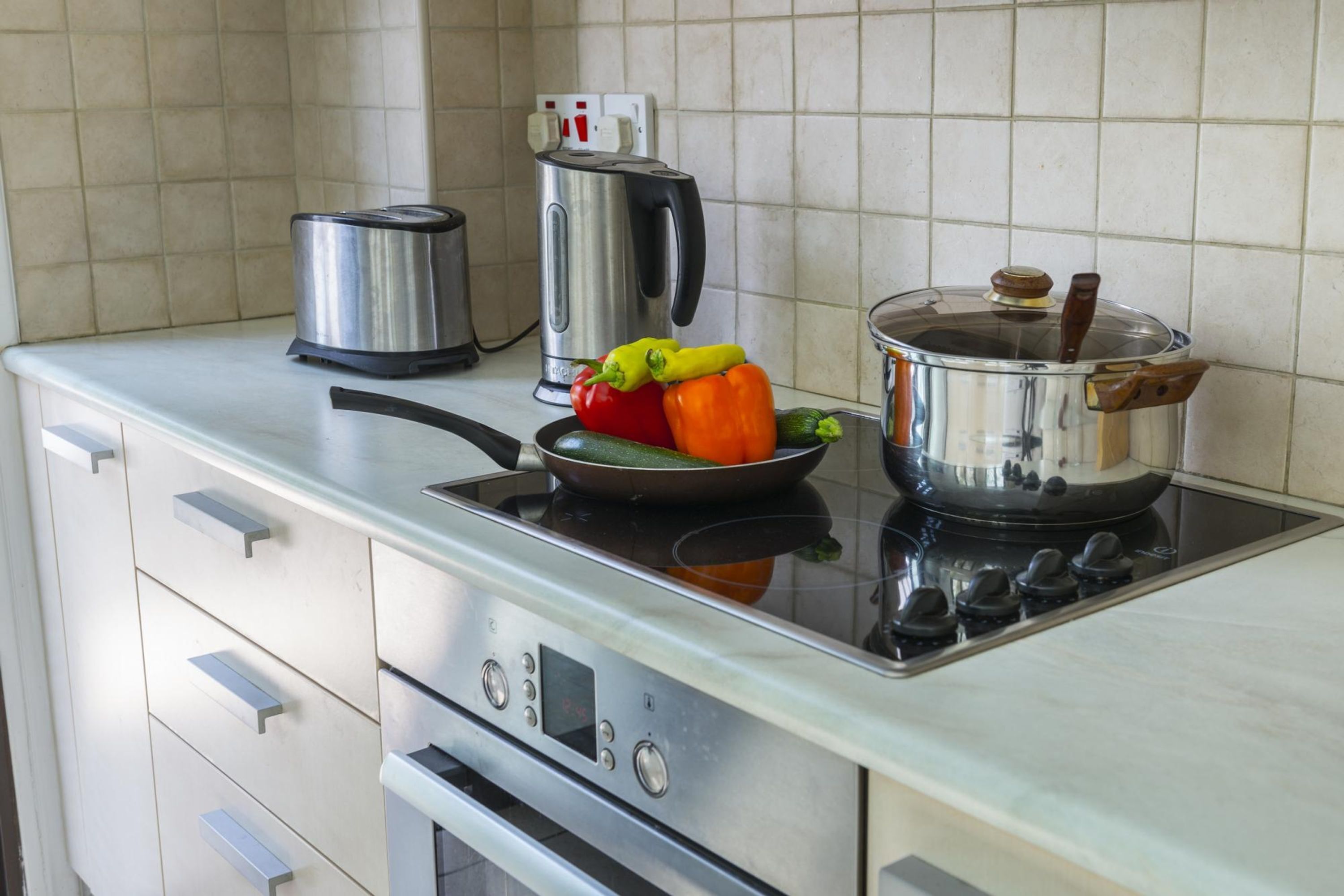 A kitchen with a stove, a kettle, a frying pan with vegetables, and a pot with a lid.