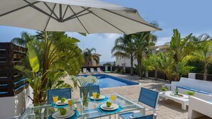 A glass table with blue placemats, white plates, and green apples, set under a large umbrella in a sunny outdoor area with a pool and palm trees. - Villa Alexa Protaras (Paralimni)