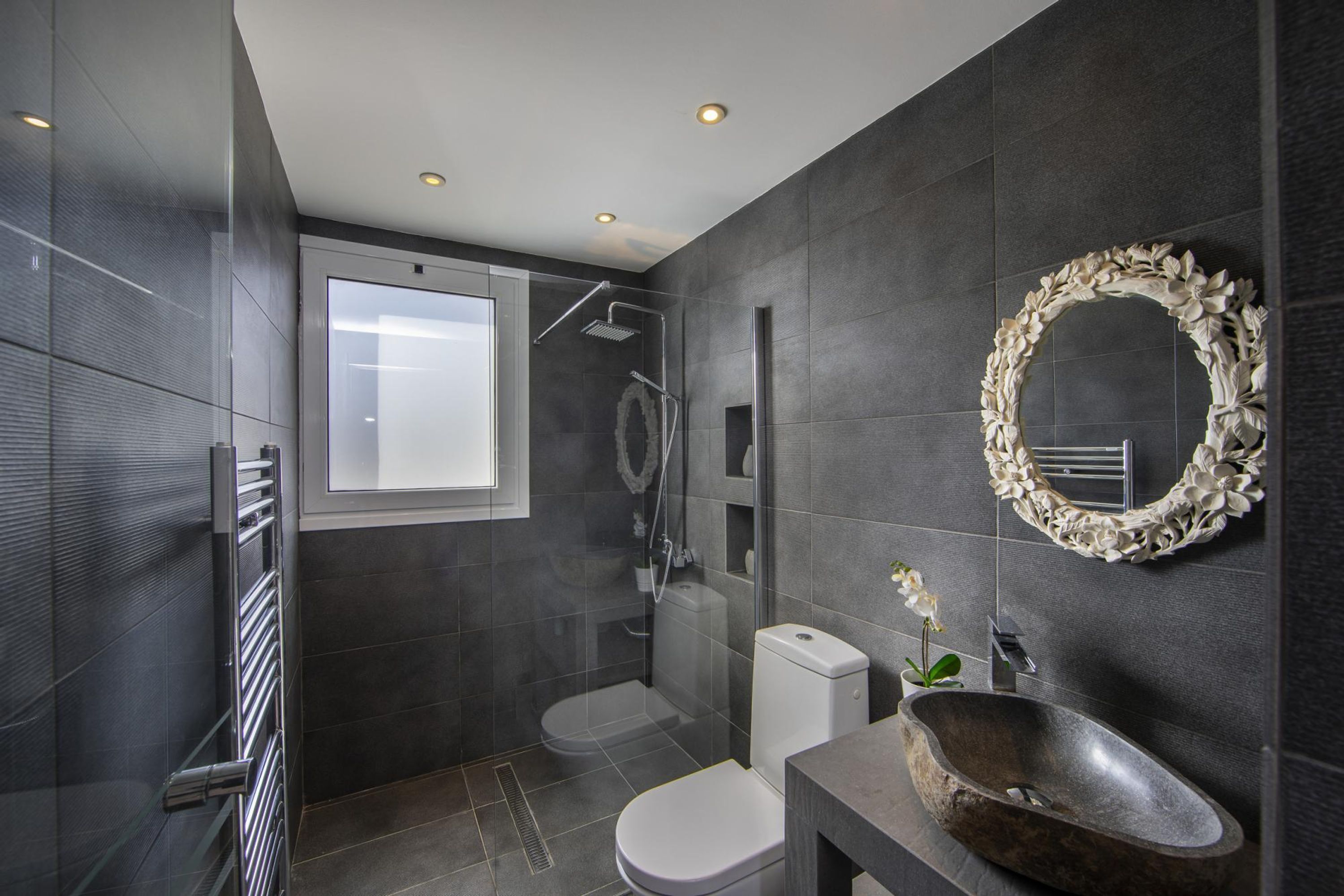A modern bathroom with a glass shower enclosure, a white toilet, a large mirror with a floral frame, and a stone sink.