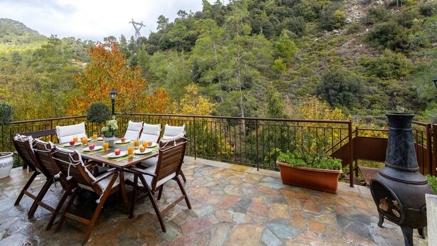 A stone patio with a table set for breakfast, surrounded by chairs, overlooking a forested hillside. - Troodos Riverside Cottage (Kato Amiantos)