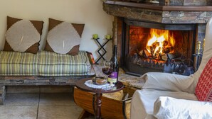A cozy living room with a stone fireplace, a sofa with patterned cushions, and a coffee table with a bottle and glasses. - Troodos Riverside Cottage (Kato Amiantos)