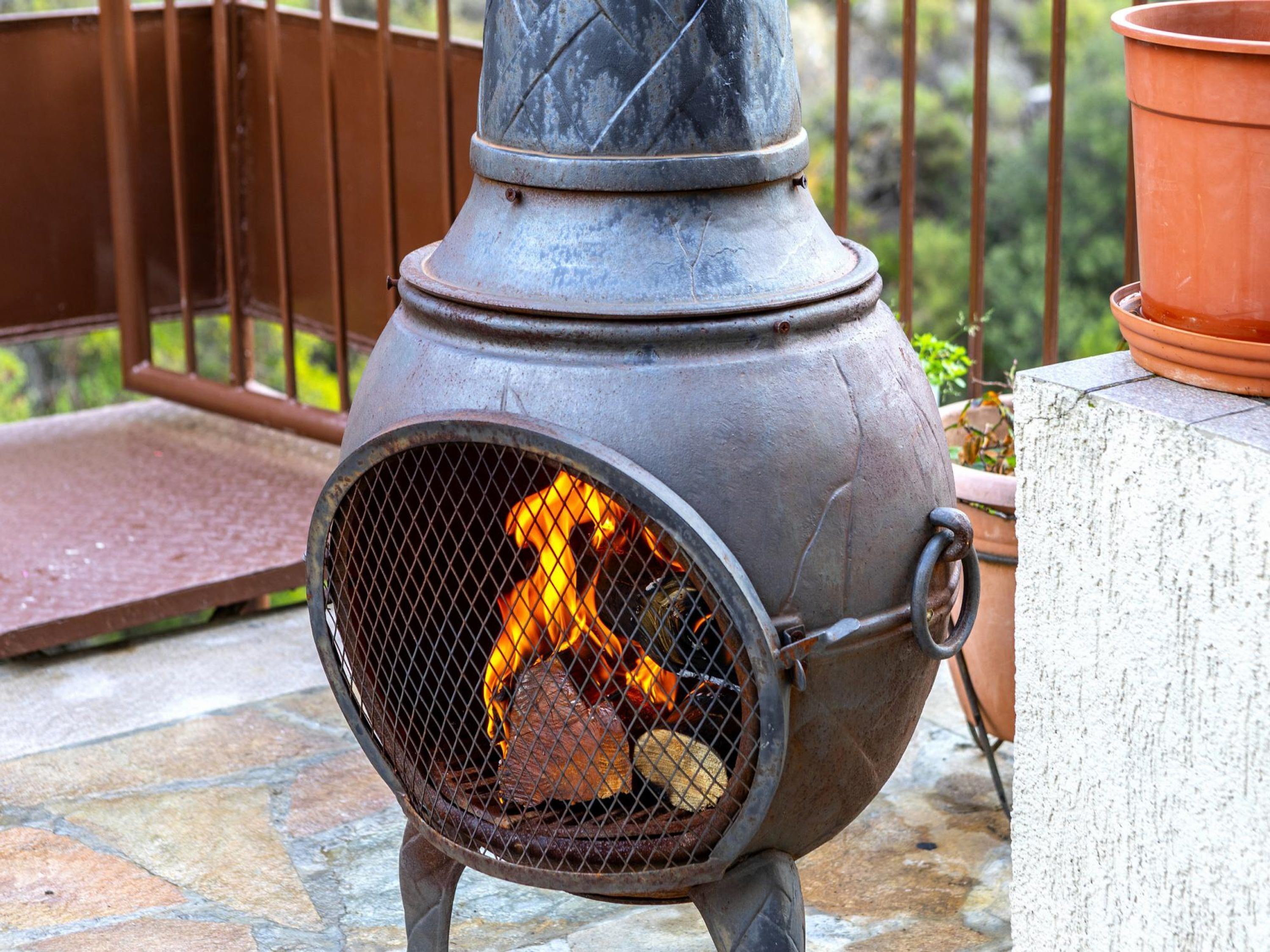 A metal outdoor stove with a fire burning inside, placed on a stone patio with a wooden bench and potted plants in the background.