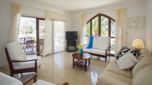 Un salon comprenant un canapé, un fauteuil et un téléviseur. On aperçoit également un coin repas avec une table et des chaises, visibles à travers une porte vitrée.