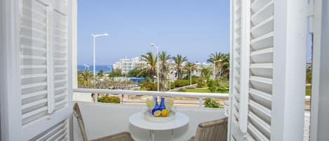 A balcony with white shutters, wicker chairs, a small round table with a white tray holding bottles and lemons, and a view of palm trees and buildings across the water.