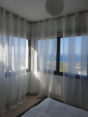 A bedroom with a bed, a window with sheer curtains, and a view of the sea and buildings.