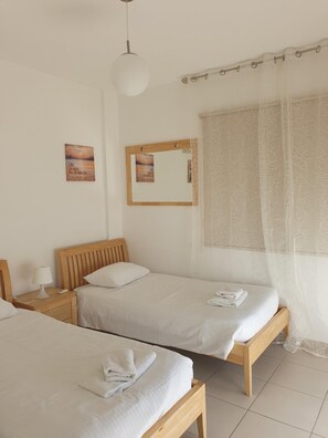A hotel room with two single beds, a bedside table with a lamp, a mirror, and a view of the sea through sheer curtains.