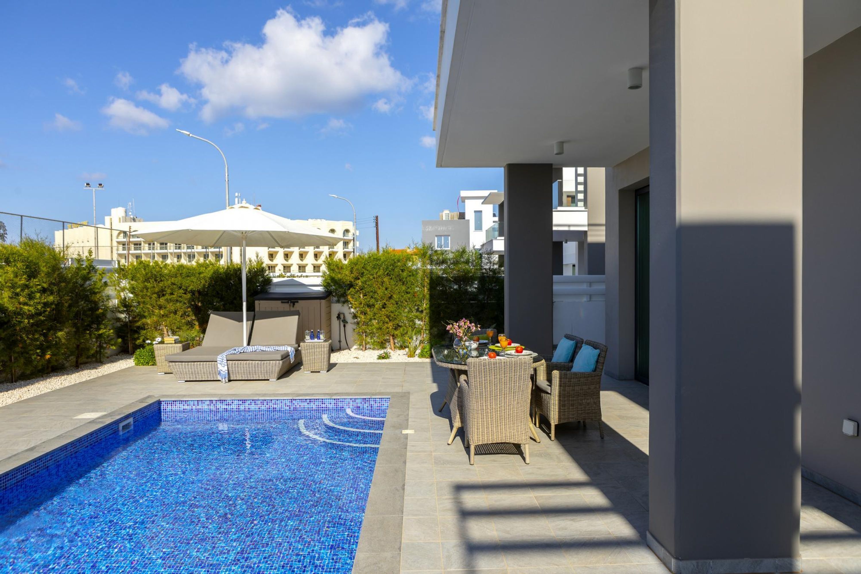 A modern outdoor pool area with lounge chairs and a dining table, surrounded by a landscaped garden and a building in the background.