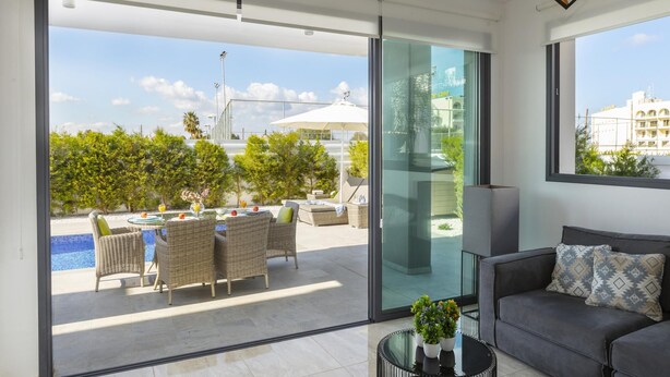 Una sala moderna con un sofá gris, un ventanal de vidrio que da a un balcón con mobiliario exterior y una piscina.